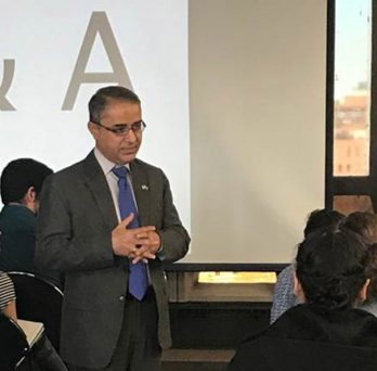 Consul General of Pakistan, Mr. Javed Ahmed Umrani, stands with fingers linked at chest in the middle of a seated audience.
                  