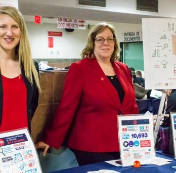 Two women dressed in red and blue and wearing UIC pins stand behind a table displaying UIC informational handouts.
                  