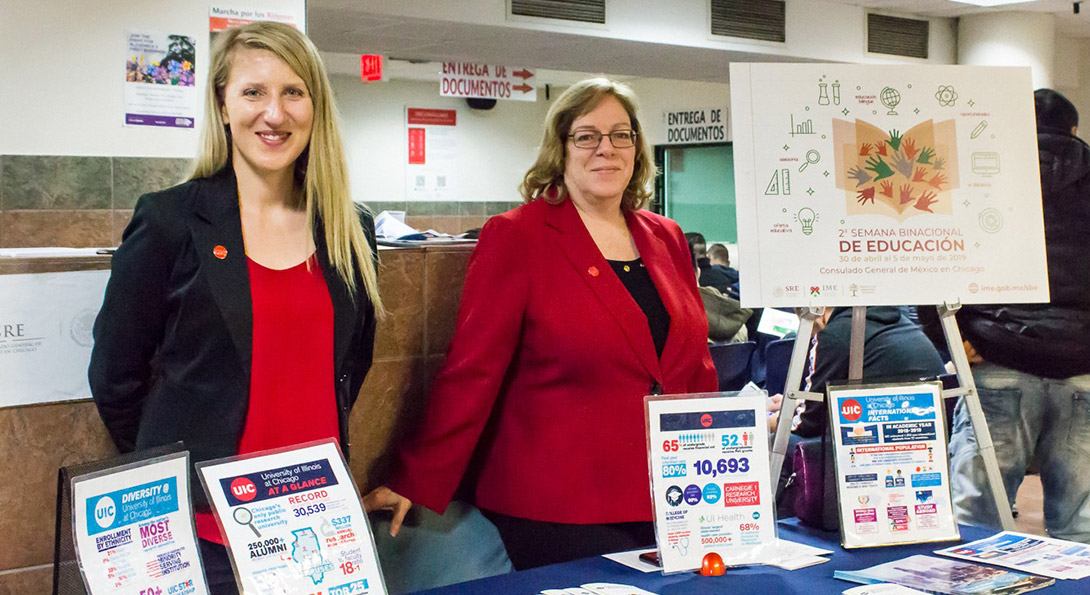 Two women dressed in red and blue and wearing UIC pins stand behind a table displaying UIC informational handouts.
