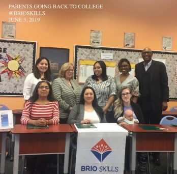 Seven women and one man pose together in a classroom. Three of the women are sitting at a table with a banner hanging off of it that says 