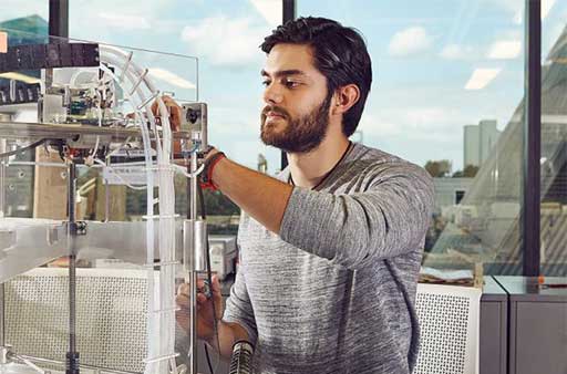 In the center of the image is a male researcher with dark hair and beard, wearing a gray long-sleeve shirt, manipulates a component of equipment in a lab with his left hand while also looking toward the equipment. In the background are floor to ceiling windows. The sky is blue with a few fluffy white clouds, and tall buildings are visible in the distance to the right of the researcher.
