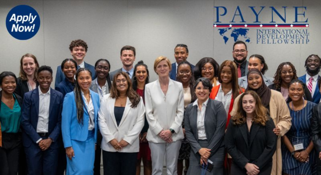 group of Payne Fellows from 2023 standing in front of the Payne Fellowship logo which is blue text on a light grey background