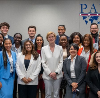 group of Payne Fellows from 2023 standing in front of the Payne Fellowship logo which is blue text on a light grey background 