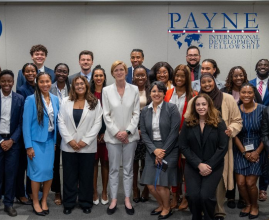 group of Payne International Fellows standing before the Payne Fellowship logo which is blue text on a grey background