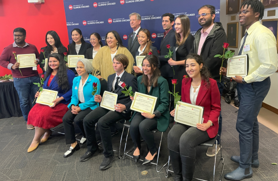 5 people seated with 11 people standing behind them, all holding a red rose and a certificate